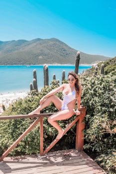 Young woman in a white swimsuit relaxing outdoors with a scenic beach and mountain backdrop.