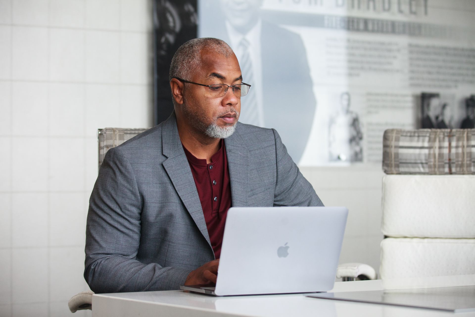 Senior adult man with gray beard working remotely on laptop indoors wearing a blazer.