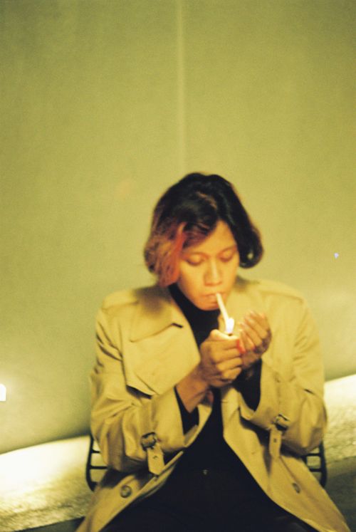 Free A blurred portrait of a person wearing a trench coat while lighting a cigarette indoors. Stock Photo