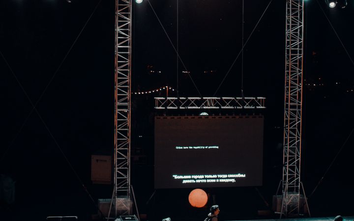 A vibrant nighttime stage setup with a crowd, lights, and a projected screen creating a lively atmosphere.