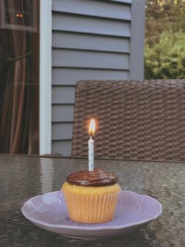 Cupcake with chocolate frosting and a lit candle on a purple plate outdoors.