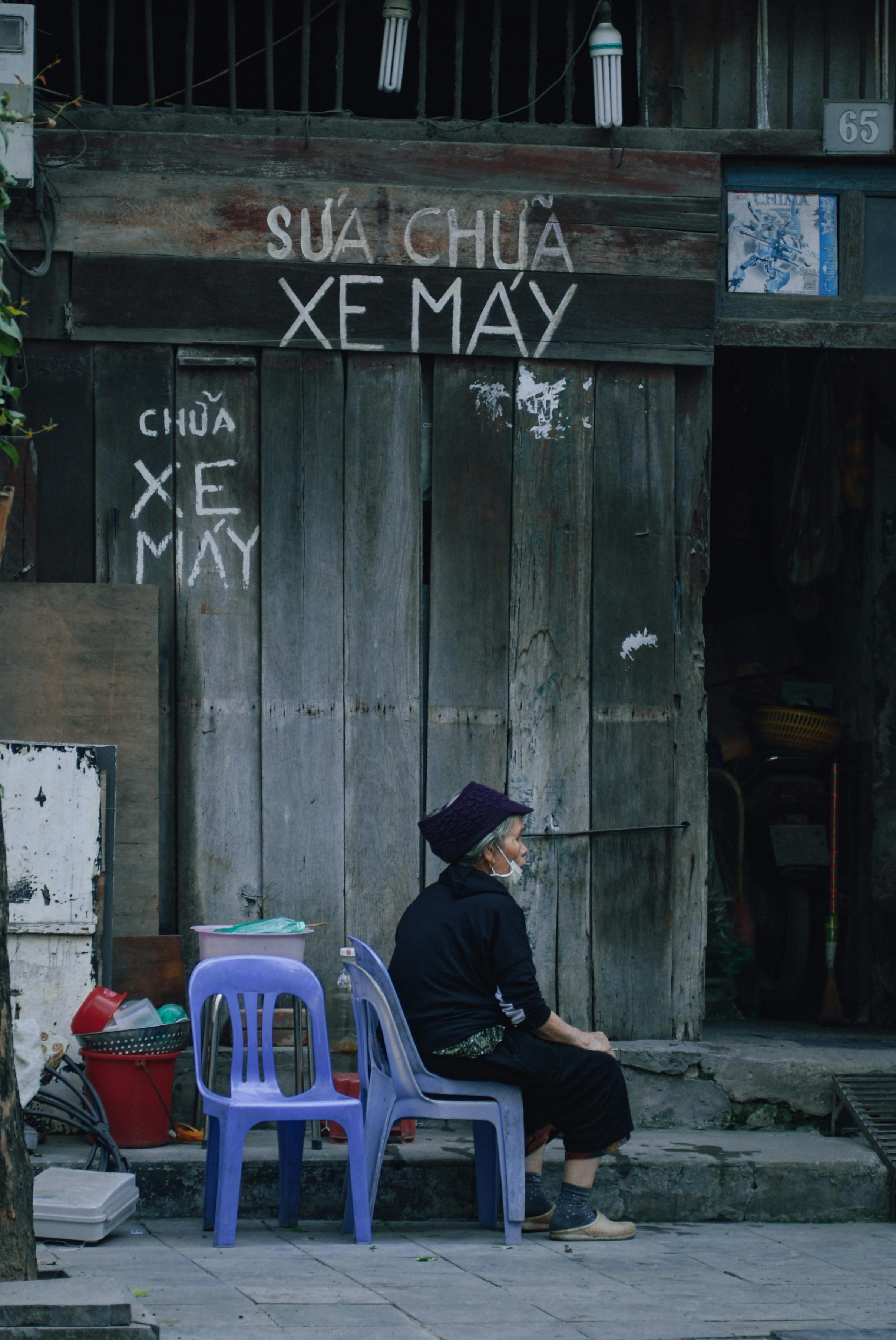 An elderly woman sits thoughtfully on a sidewalk in Hanoi, reflecting everyday life.