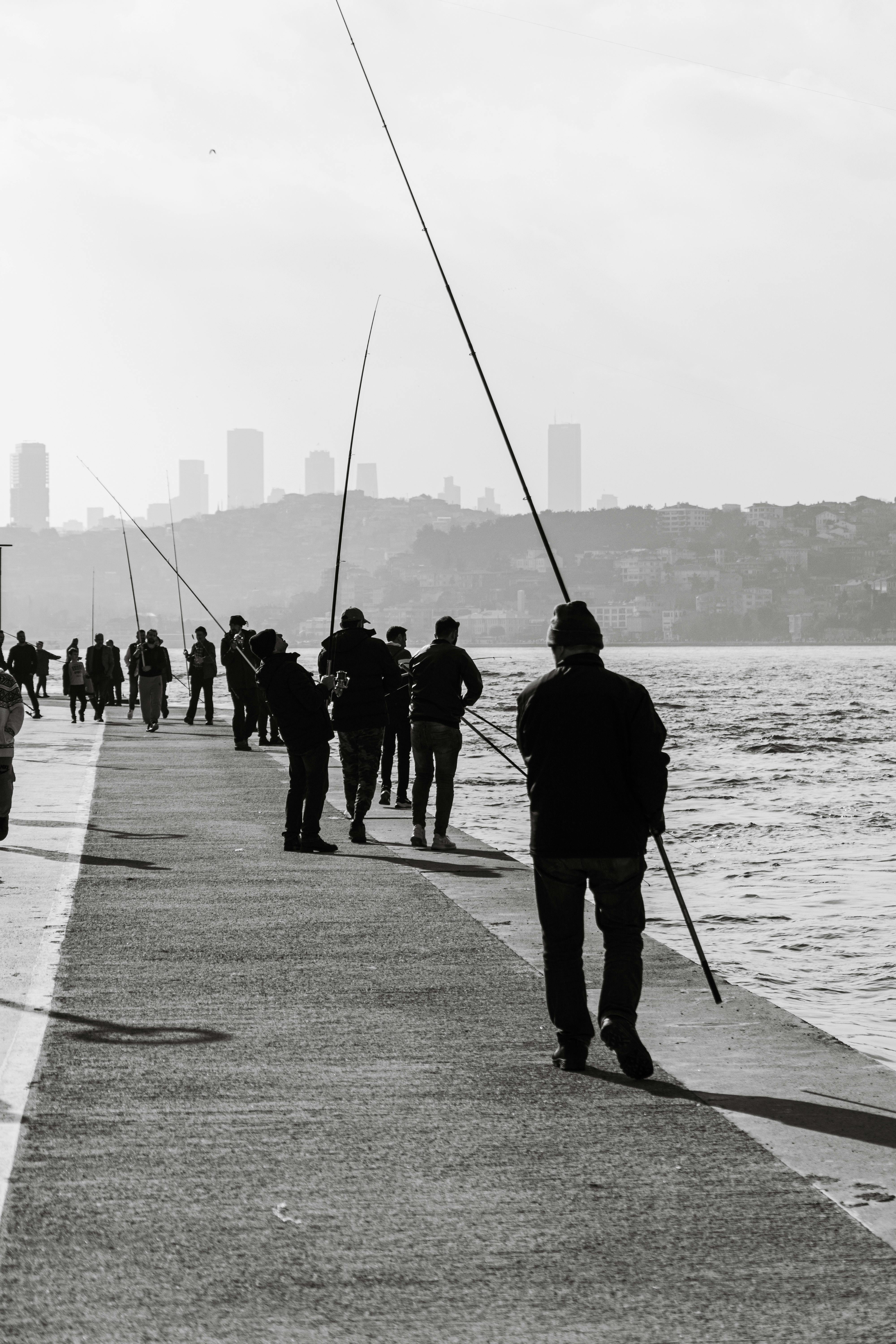 Black and white photo of people fishing on an urban pier, city skyline visible.