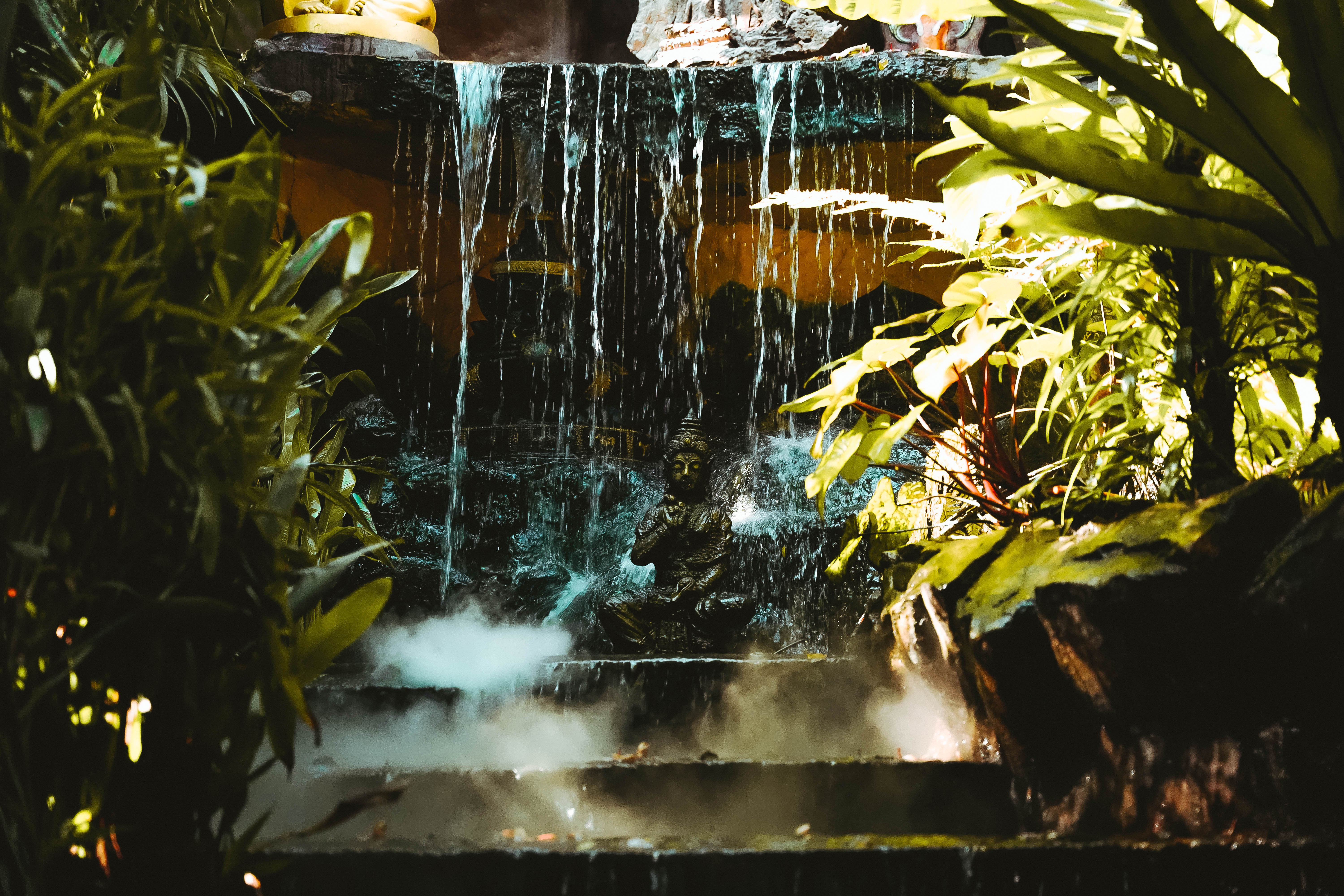 A serene Buddha statue surrounded by lush foliage and a cascading waterfall in Bangkok, Thailand.