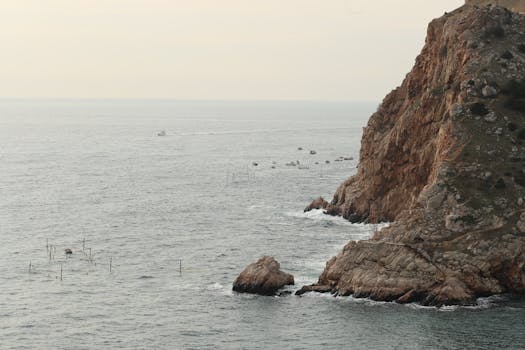 Aerial view of a rugged coastline with boats navigating the calm sea under a white sky.