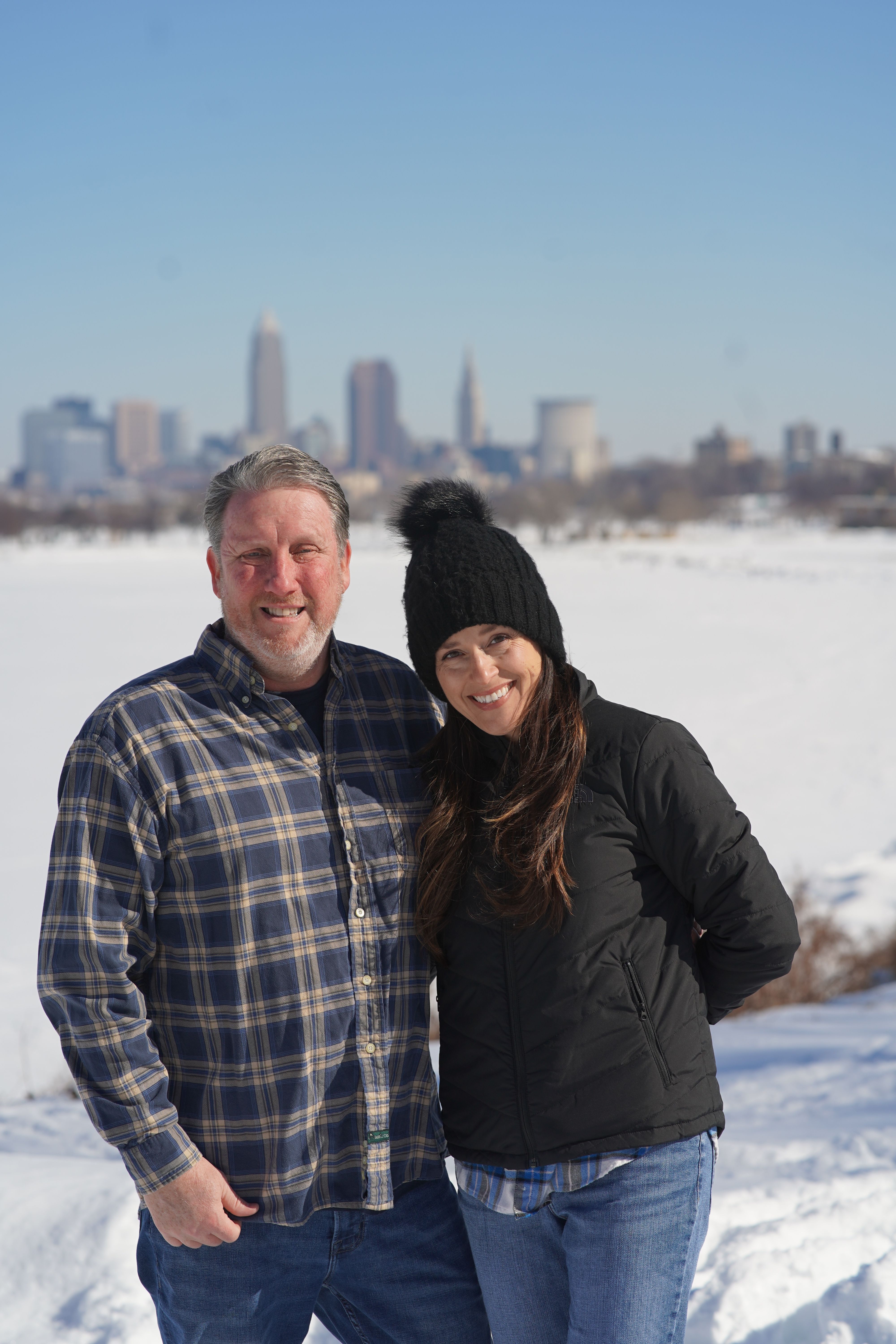 Couple smiling in snowy park with city skyline in background.