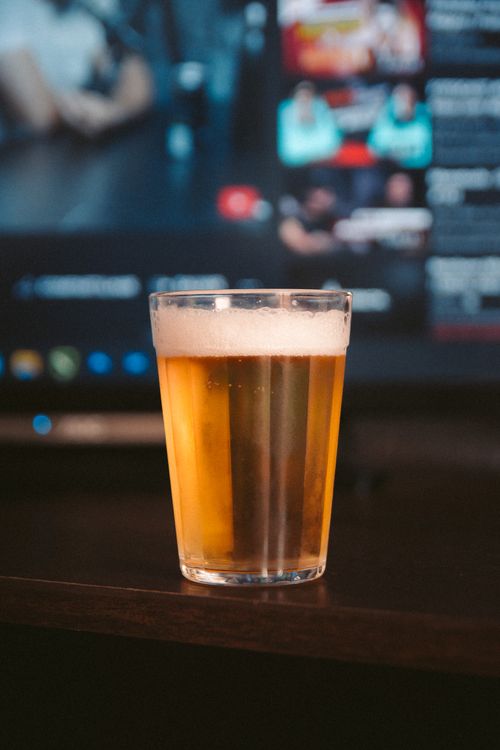 Free Glass of beer with frothy head placed indoors near a TV screen, evoking a relaxed atmosphere. Stock Photo