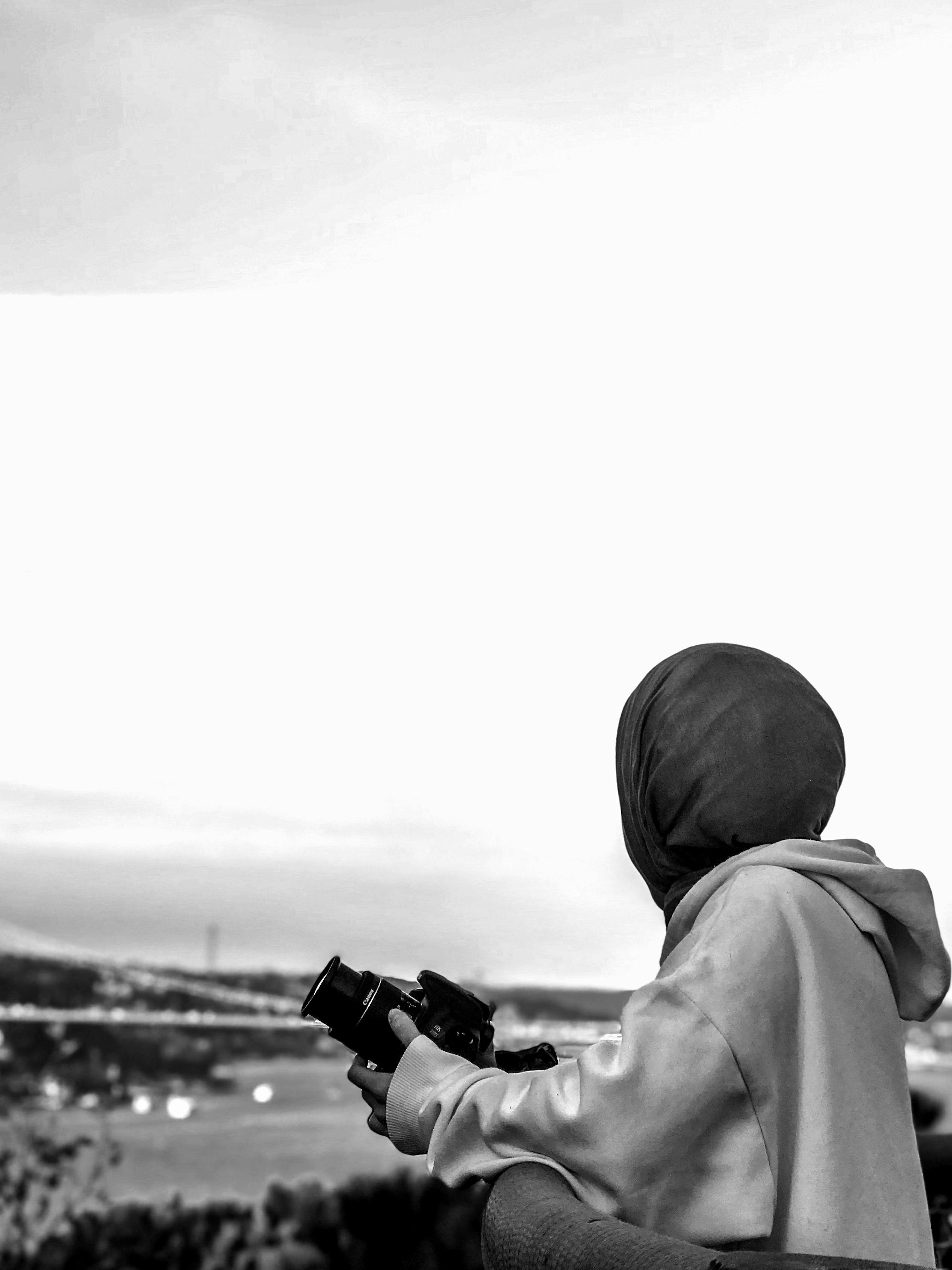 A woman in a headscarf holding a camera, gazing over a landscape in black and white.