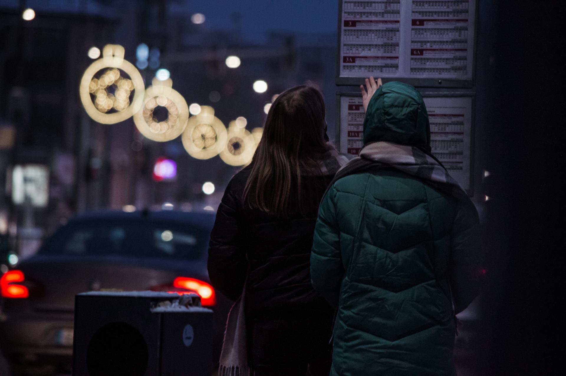 Two people check a bus schedule on a winter night, illuminated by street lights and festive decorations.
