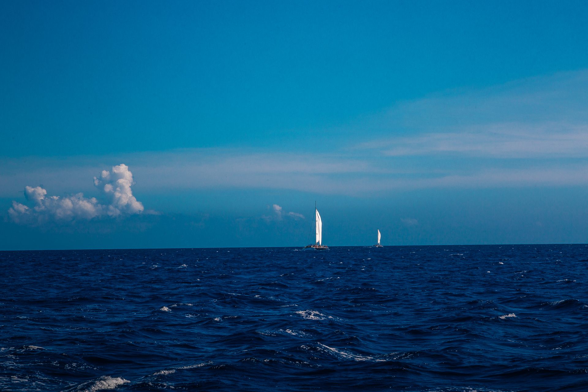 Scenic view of sailboats navigating the deep blue ocean with fluffy white clouds and clear skies.