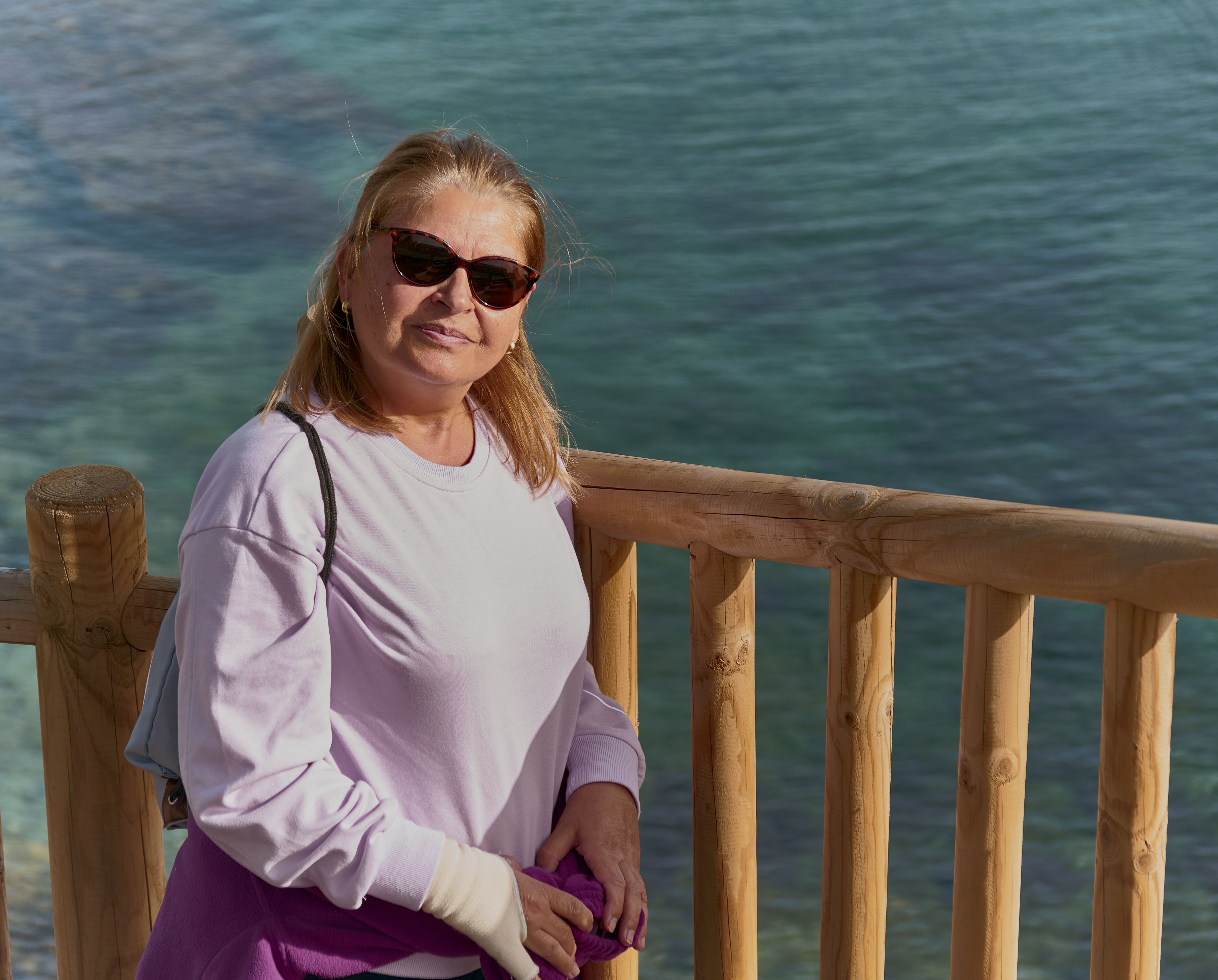 Senior woman in sunglasses at a wooden railing by the sea on a sunny day.