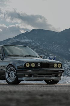 Classic BMW E30 parked with a stunning mountain backdrop under a cloudy sky.