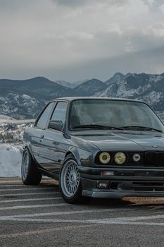 A classic black BMW E30 car parked with scenic snow-capped mountains in the background, emitting a vintage allure.