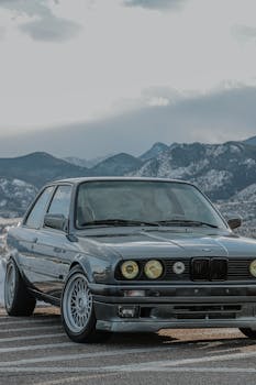 Classic BMW with headlights on, parked on a road with mountain backdrop under cloudy sky.