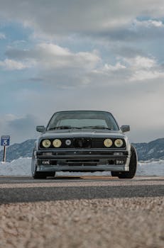 Front view of a classic BMW E30 parked on a mountain road under a blue sky.