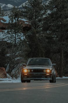A vintage BMW E30 car driving through a scenic snowy mountain road surrounded by trees.