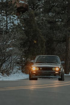 Captivating shot of a vintage BMW E30 driving through a snow-covered forest road under low-light conditions.