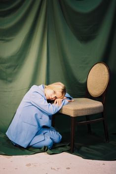 Artistic studio portrait of a blonde woman kneeling in a blue suit next to a chair and green curtain.