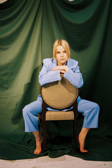 A woman with blond hair in a blue suit sits barefoot on a chair against a green backdrop.