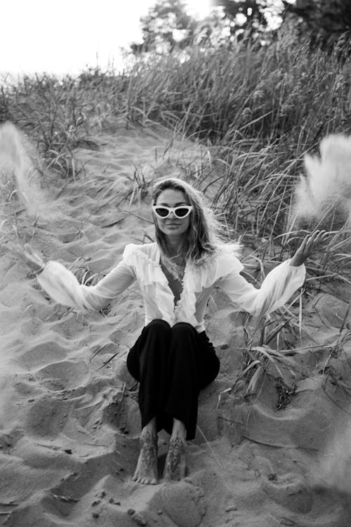 A Grayscale Photo of a Woman Sitting on the Sand Near the Grass Field