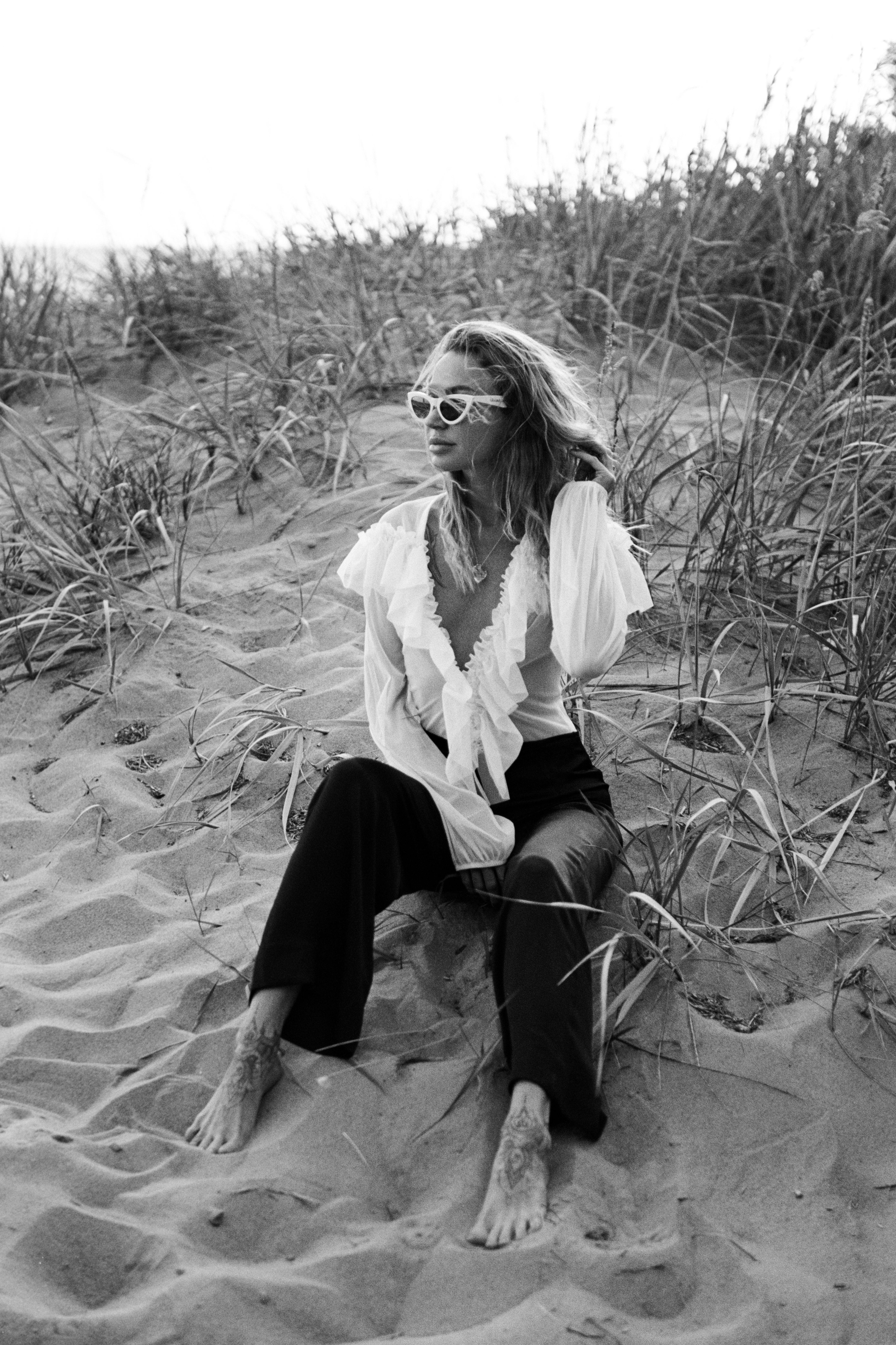 Black and white photo of a stylish woman sitting on a sandy beach wearing sunglasses and a ruffled shirt.