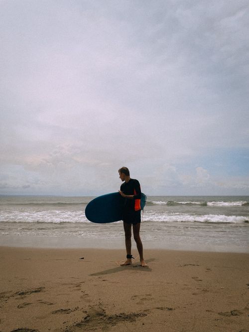 Free A surfer standing on the sandy beach of Bali, Indonesia, holding a surfboard and looking towards the ocean. Stock Photo