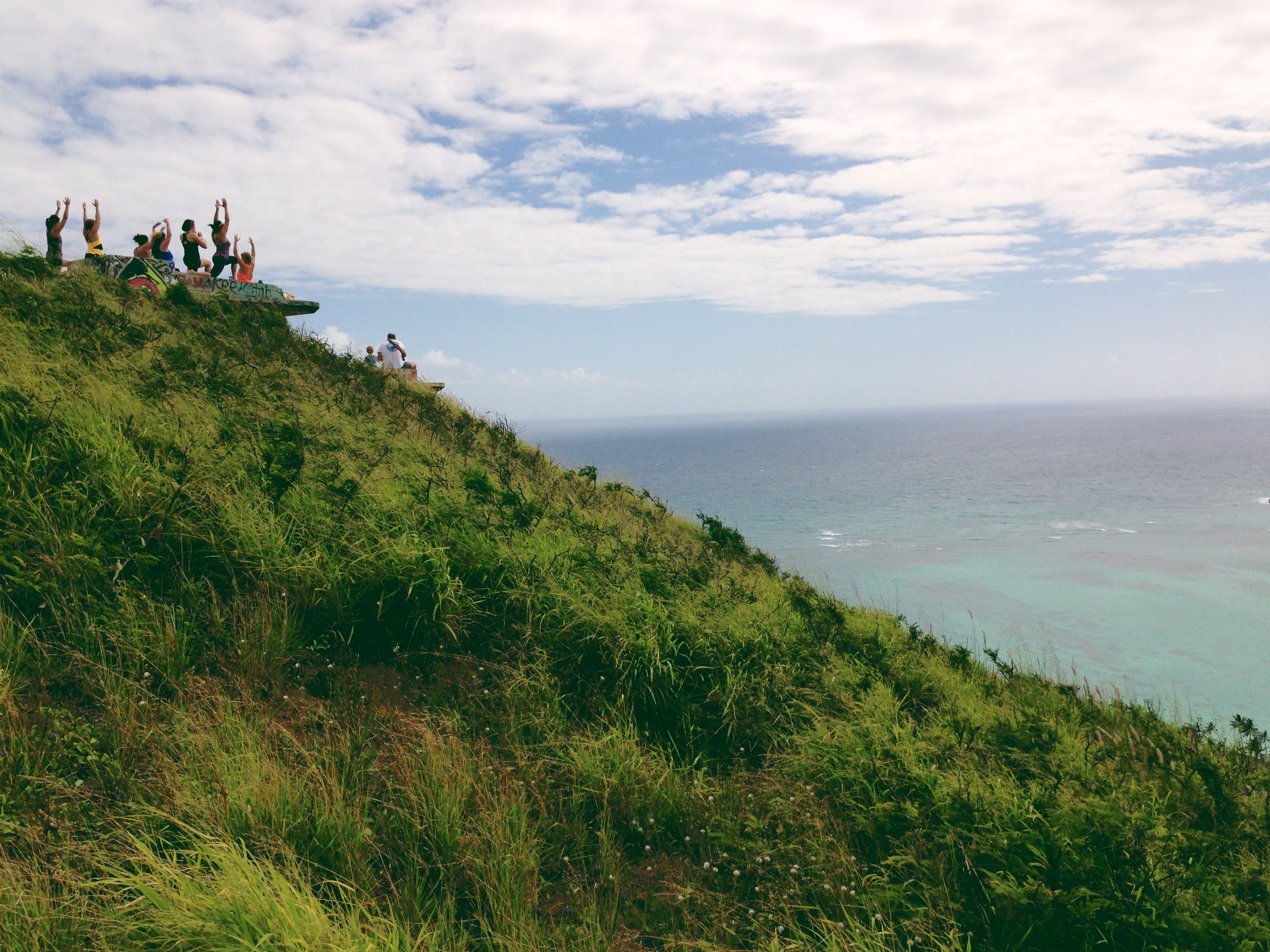 Group practicing yoga atop lush green hill with breathtaking Kailua ocean views on a sunny day.