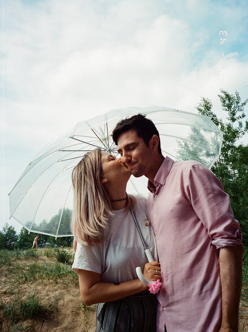 A Woman in White Shirt Holding an Umbrella while Kissing a Man in the Cheeks