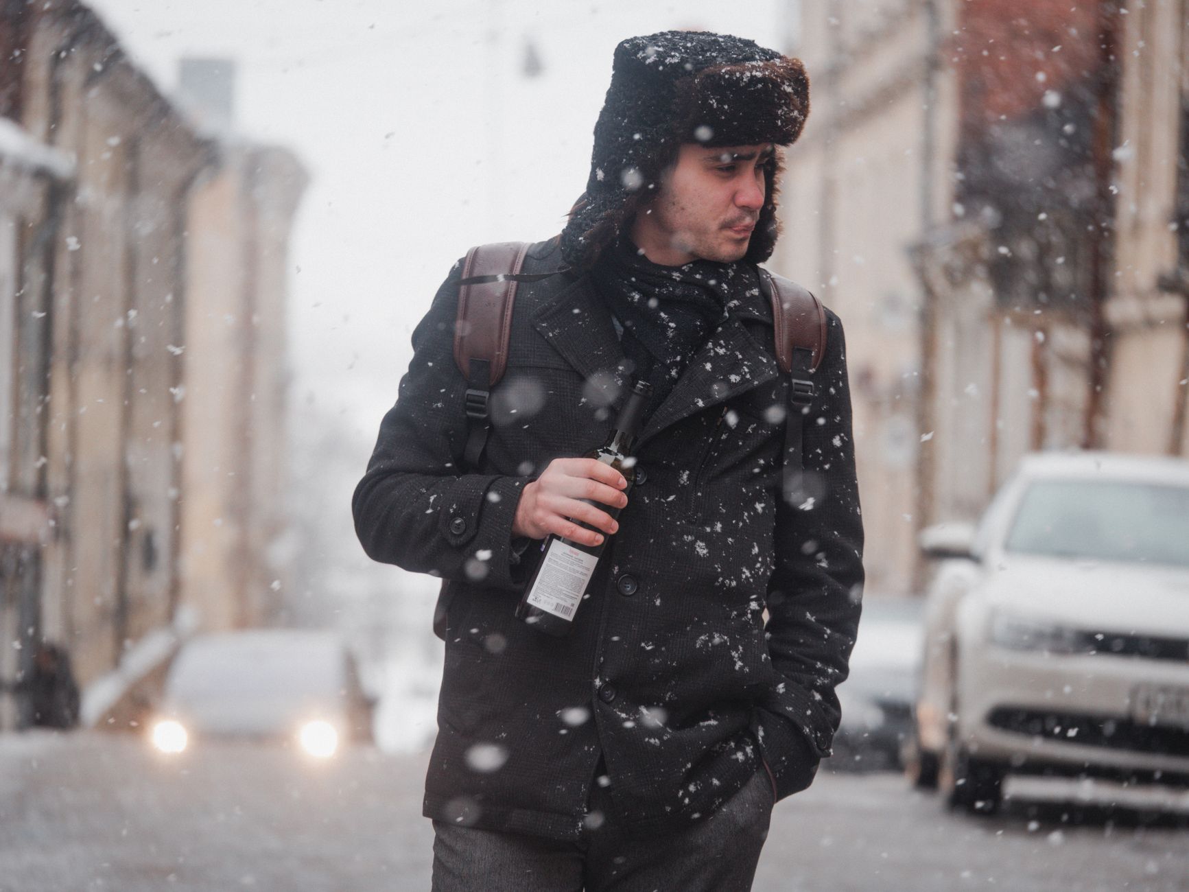 A man in winter clothing holding a bottle, walking on a snowy city street.