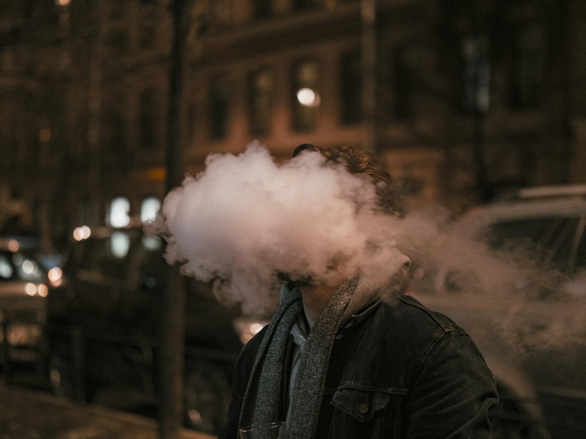 A man exhales a thick cloud of vapor on a city street at night, creating an artistic and dramatic effect.