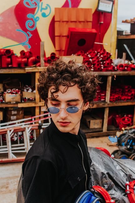 Trendy young man with curly hair wearing sunglasses, standing against colorful industrial backdrop.