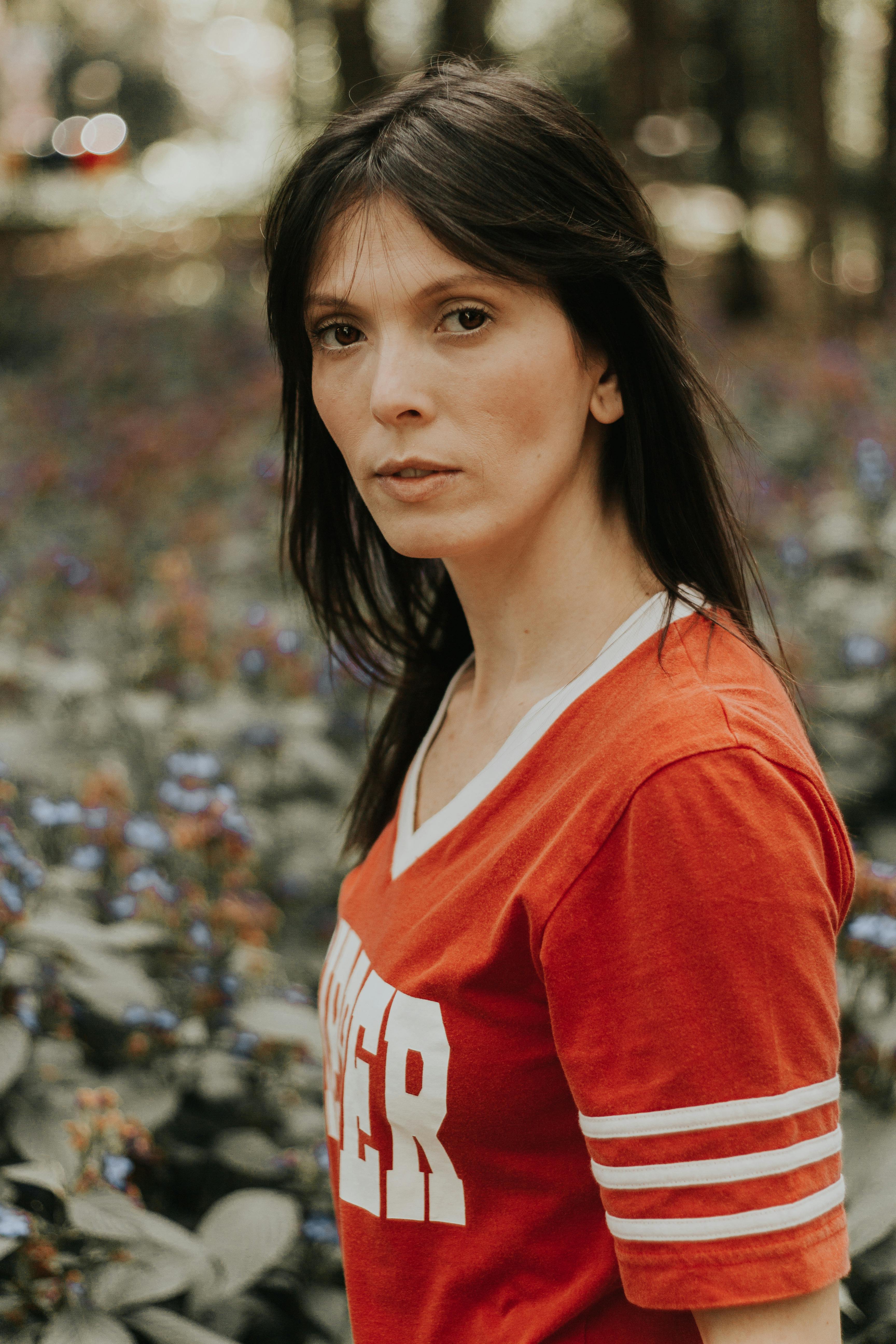 Portrait of a woman standing outdoors in a red shirt amidst lush foliage.