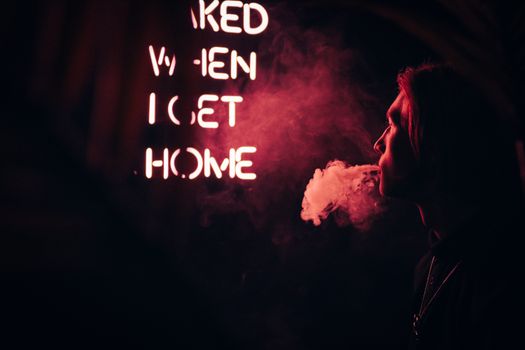 A side view of a person exhaling smoke near neon lights in a dark urban setting.