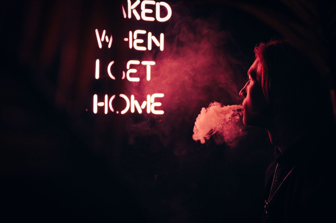 a side view of a person exhaling smoke near neon lights in a dark urba