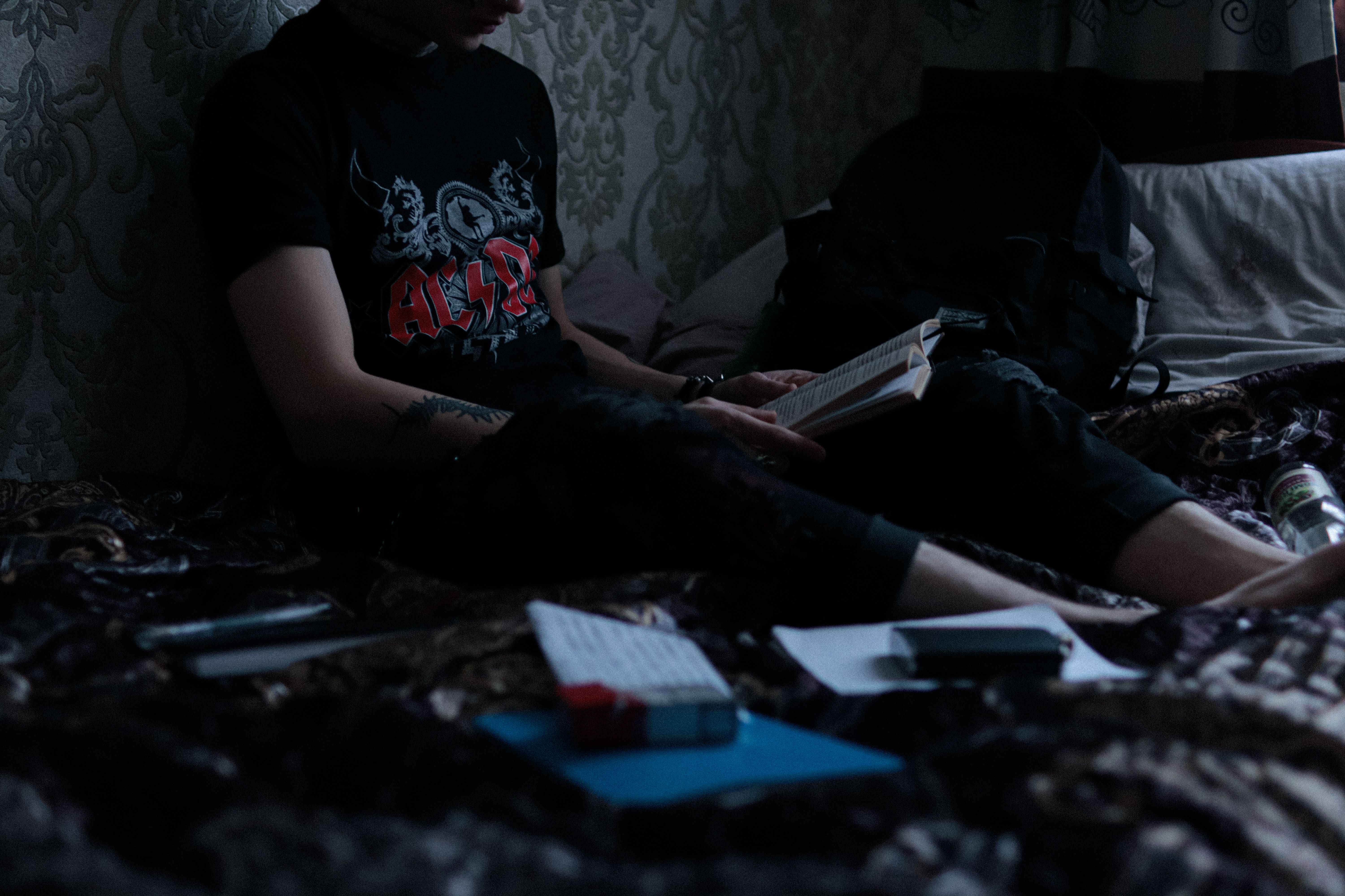A young person sits on a bed, reading a book in a dimly lit room with casual surroundings.