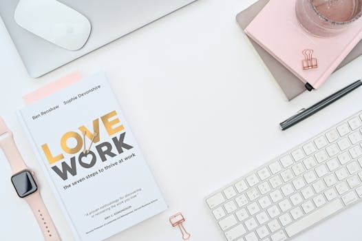 A clean, modern workspace layout featuring a book, Apple Watch, and keyboard on a white background.