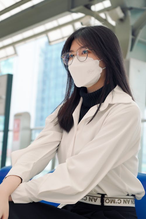 A Low Angle Shot of a Woman in White Long Sleeves Sitting while Wearing Face Mask and Eyeglasses