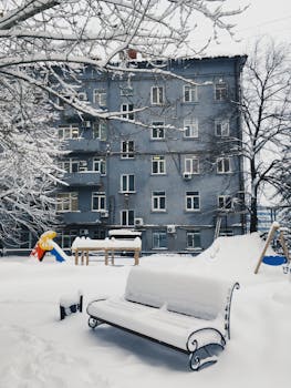 A snow-laden playground and bench outside an apartment building during winter.