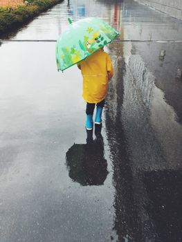 A child in a yellow raincoat walks on a wet street holding a colorful umbrella reflecting in puddles.