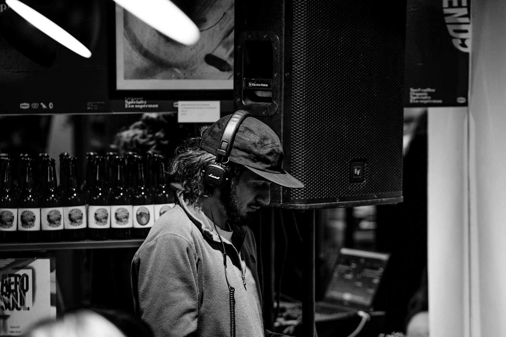 Black and white photo of a DJ with headphones, bottles, and speaker in an indoor setting.