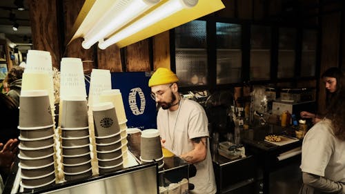 Free A Man in White Shirt and Yellow Beanie Standing Near the Counter with Disposable Cups Stock Photo