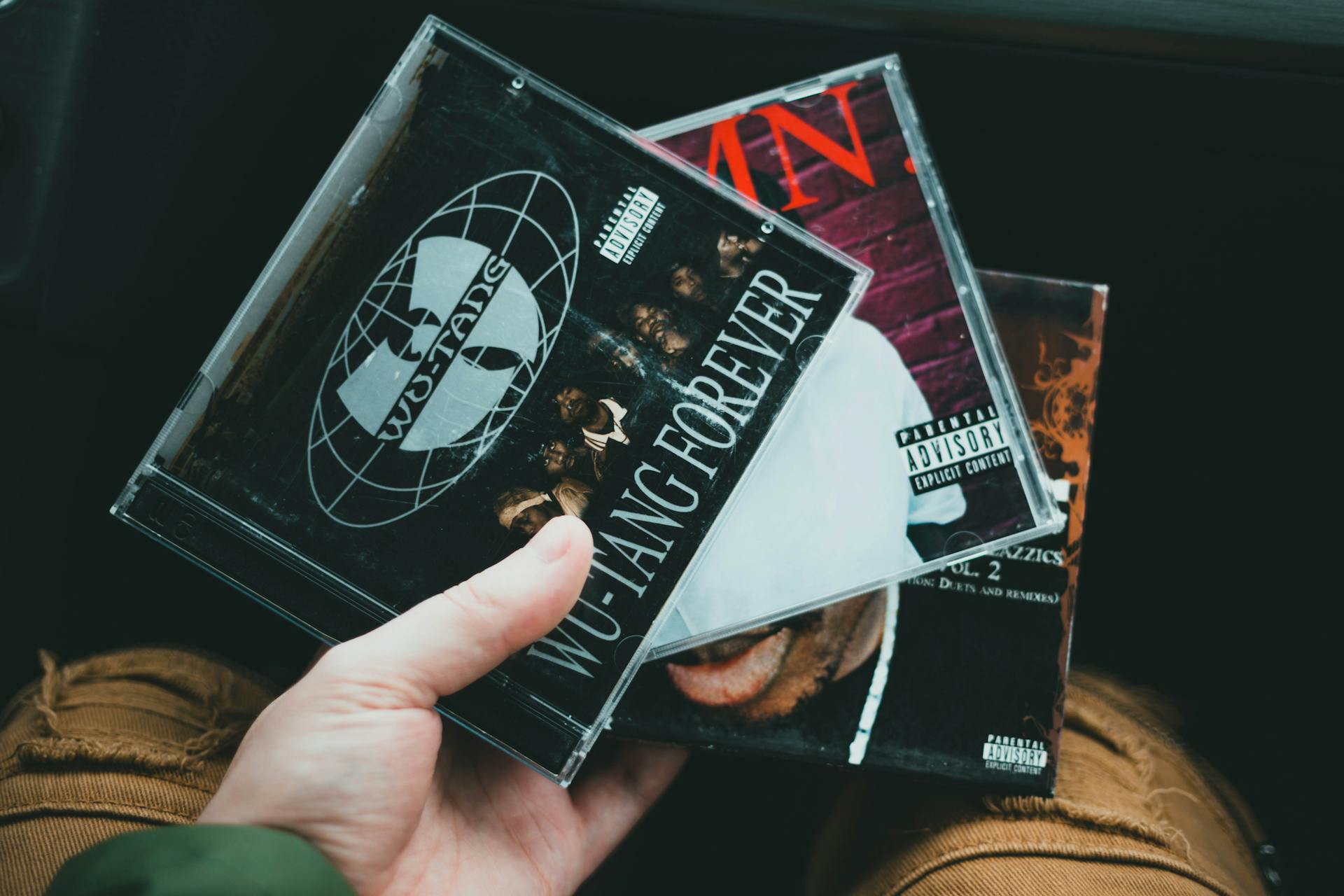 A hand holding a collection of classic hip hop CDs showcasing iconic album covers.