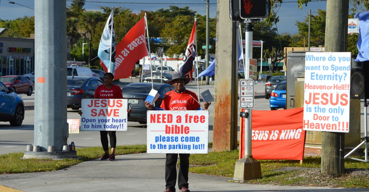 Two individuals holding religious signs on a city sidewalk, conveying Christianity messages.