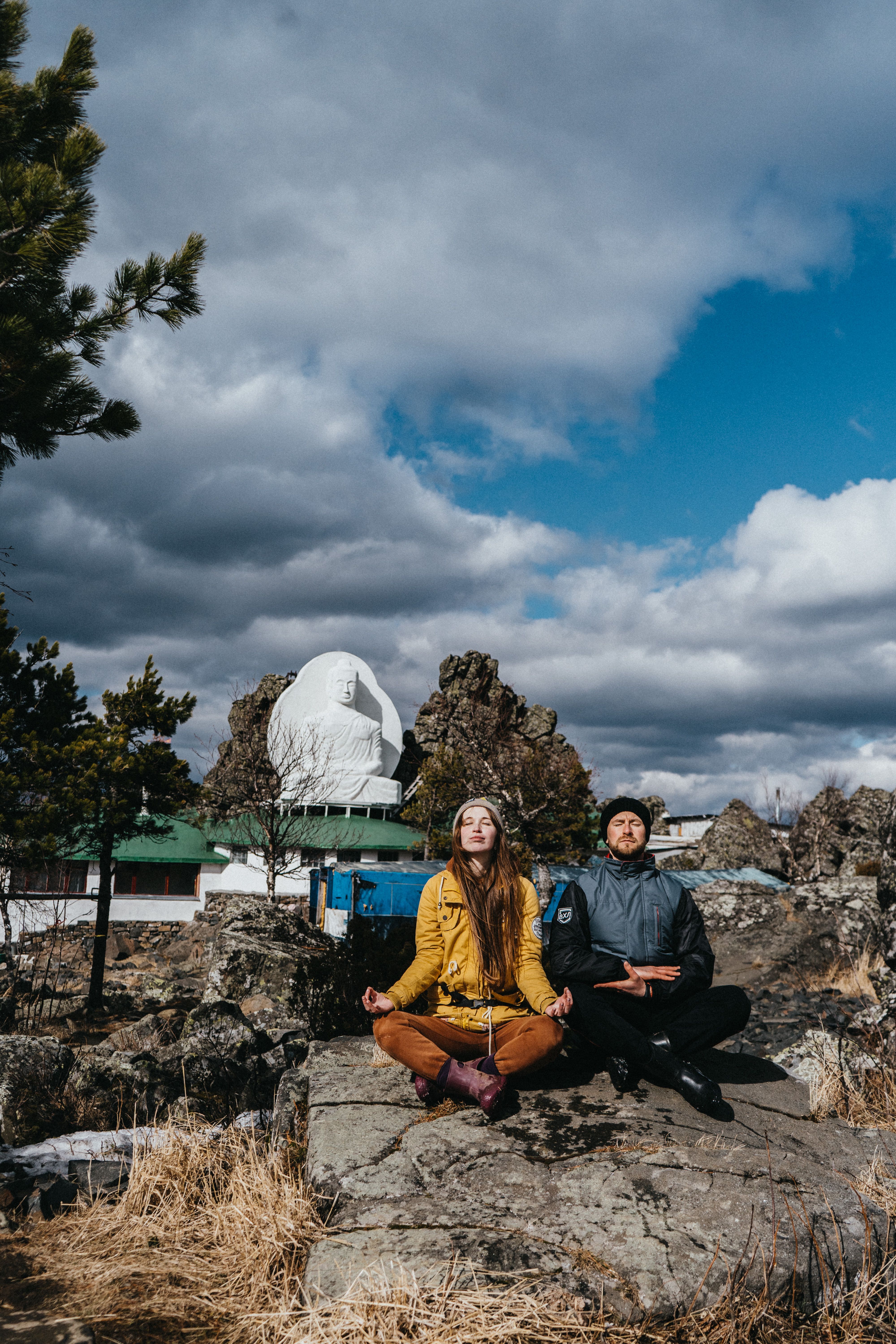A couple sitting in meditation pose outdoors near a large Buddha statue. Calm and tranquil setting.