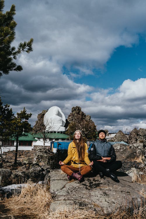 A Man and a Woma Doing Yoga while Sitting on Rock Under White Clouds and Blue Sky