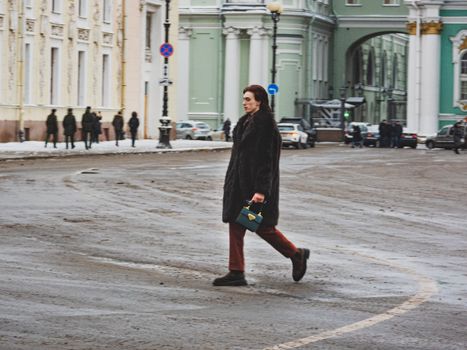 Stylish man with fur coat and purse walking through a wintry city street.