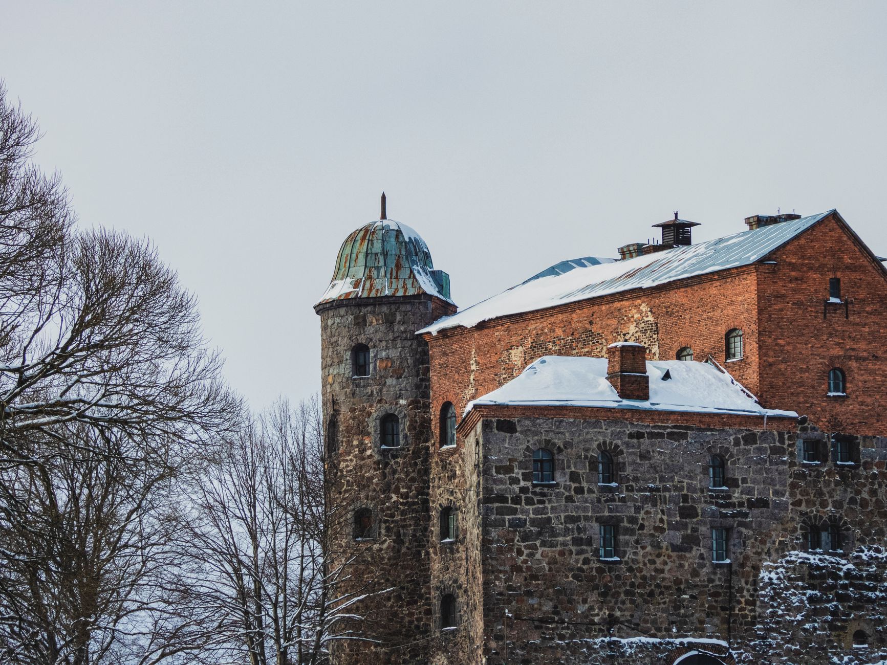 A beautiful old castle with red brick and stone walls covered in snow, surrounded by barren trees in winter.