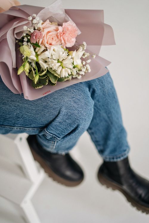 Free Close-up of a person holding a delicate floral bouquet, wearing jeans and boots, seated on a white chair. Stock Photo