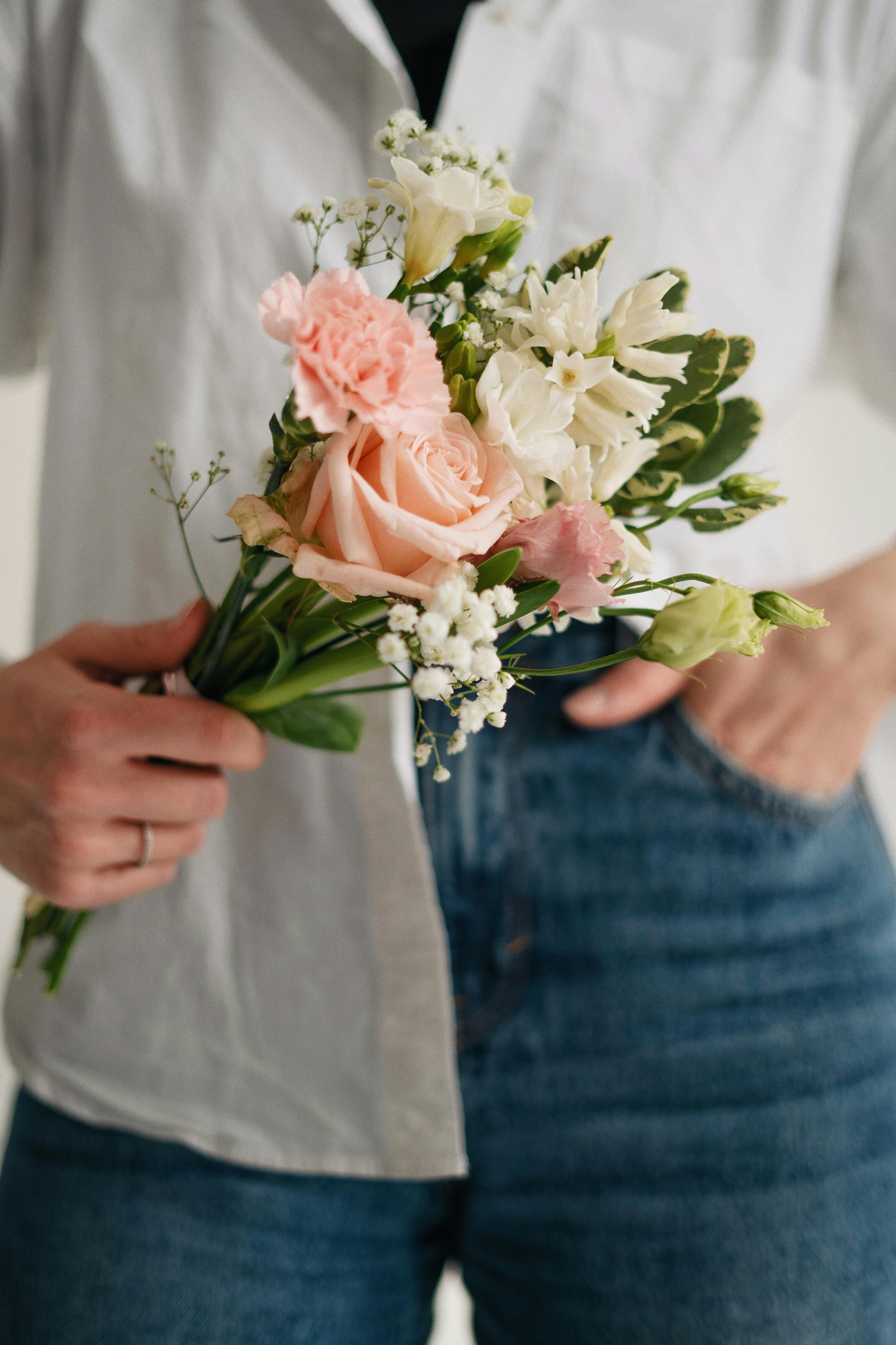 A close-up of a person holding a delicate bouquet of pink roses and other flowers against a white shirt.