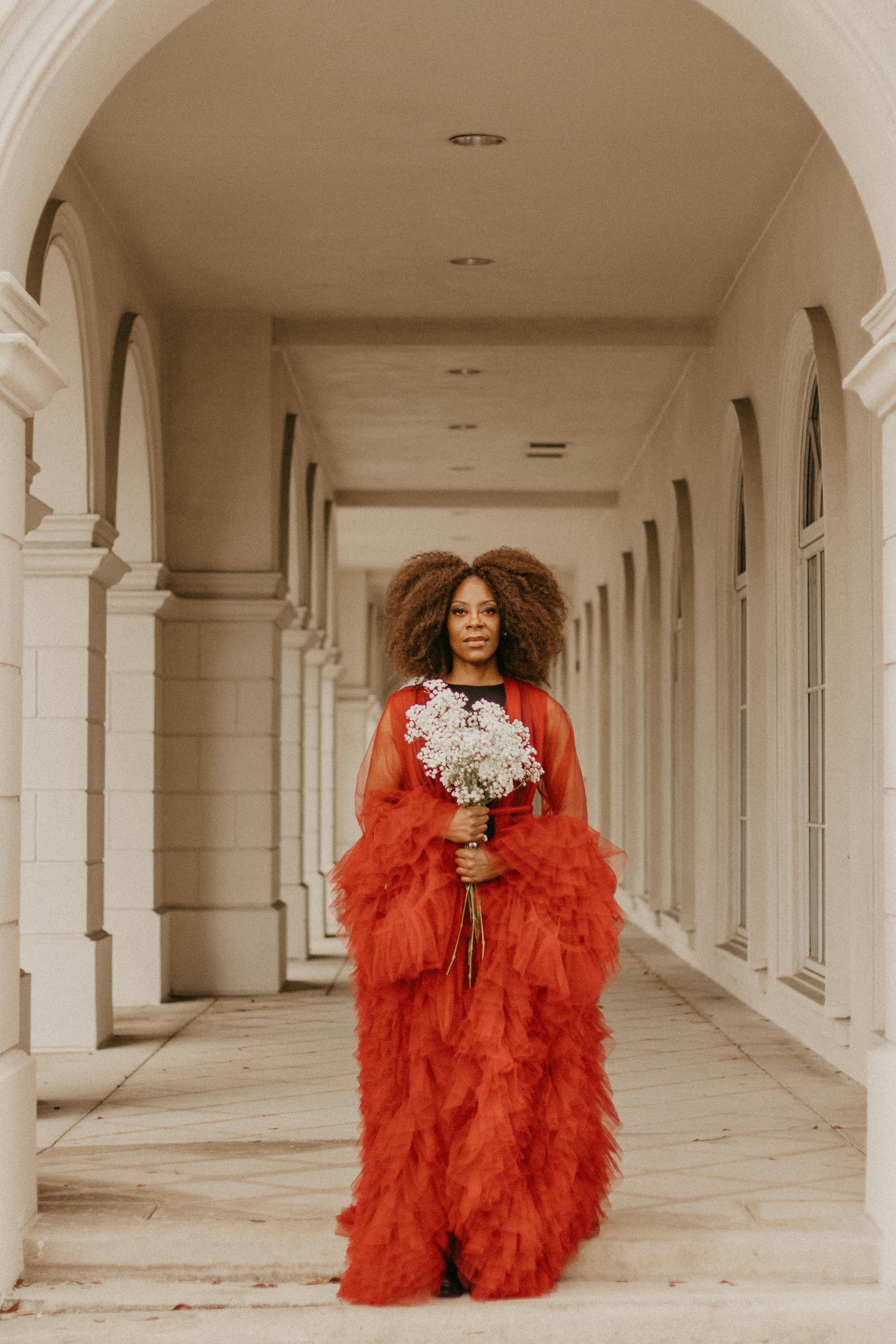 A stylish woman in a vibrant red gown holding a bouquet in an elegant corridor.